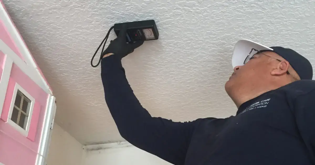 Technician holds a pinless moisture meter flat against a textured ceiling near a stained ceiling-wall joint