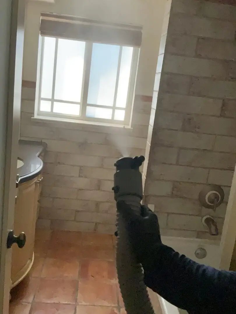 Gloved hand fogging a tiled bathroom with a sanitizing mist.