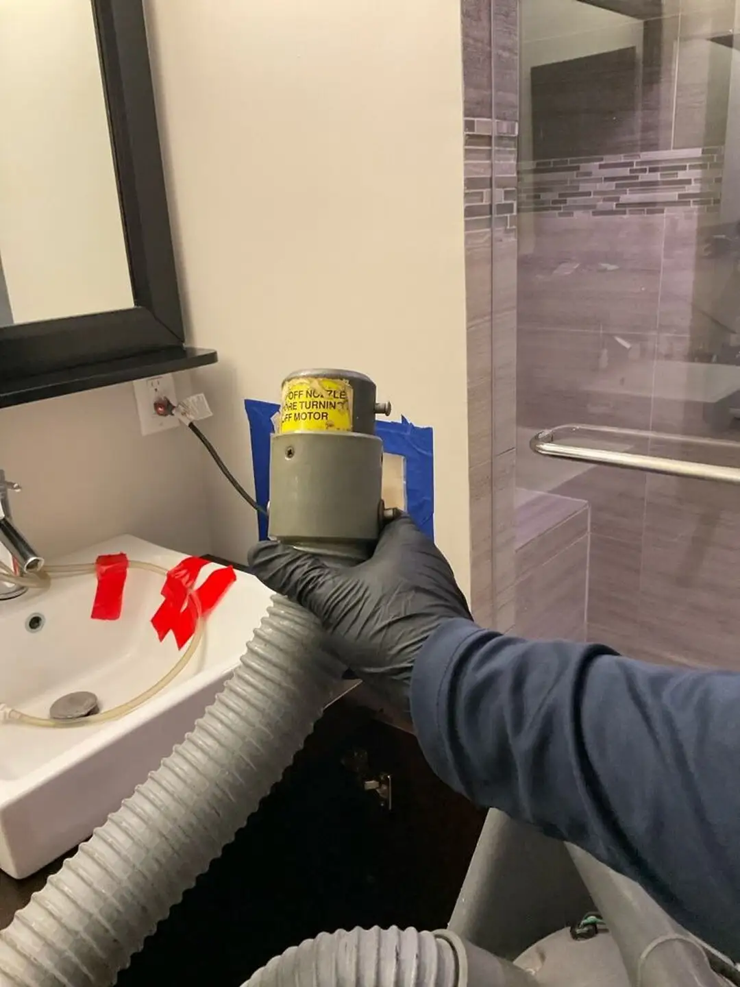 Gloved hand holds a gray extraction nozzle in a bathroom beside a taped wall opening.