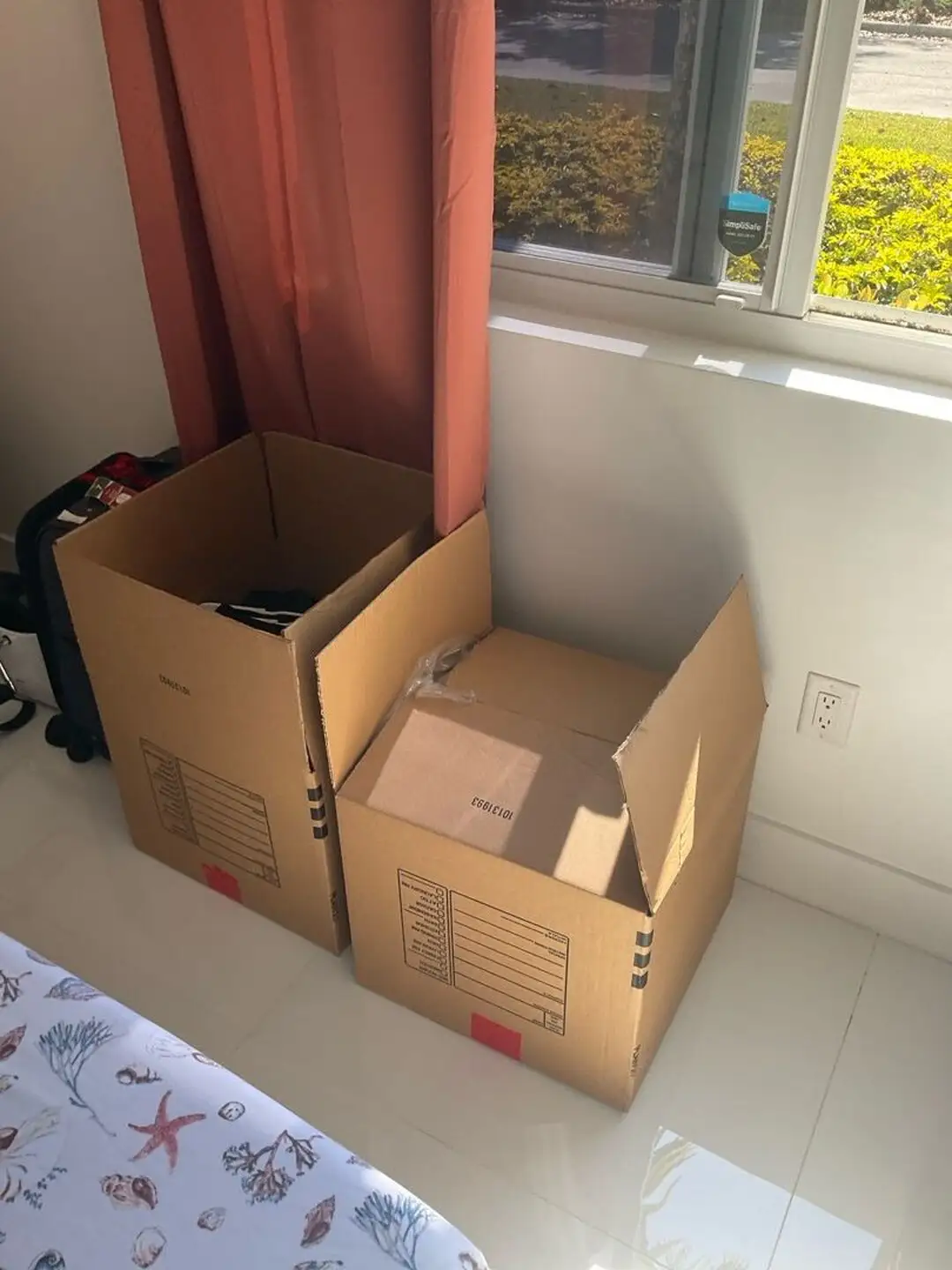 Two cardboard pack-out boxes on a tiled bedroom floor beside a window and bed.