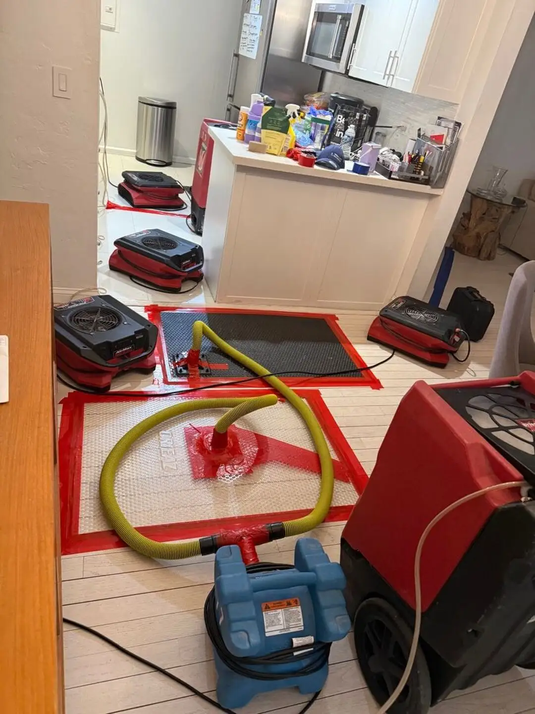 Hardwood-floor drying mats taped down with yellow hoses, air movers, and a dehumidifier in a kitchen.