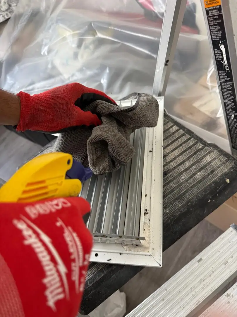 Gloved hands wipe a metal HVAC register on a stepladder with a cloth and spray bottle.