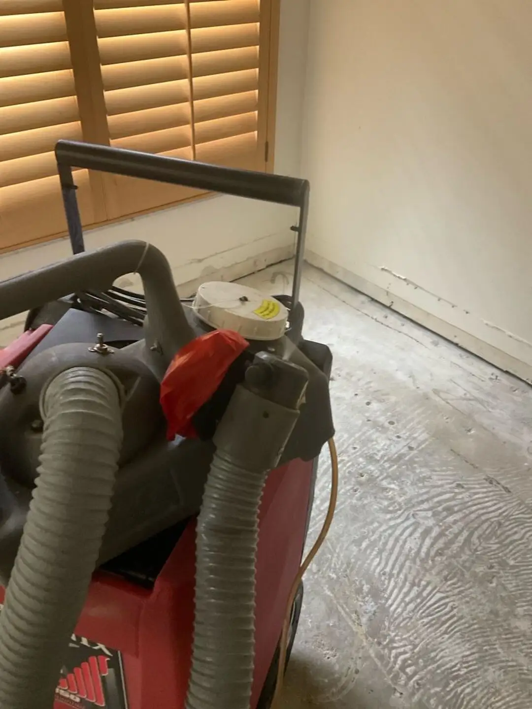 Red industrial dehumidifier with gray hoses in a room with baseboards removed and plantation shutters.