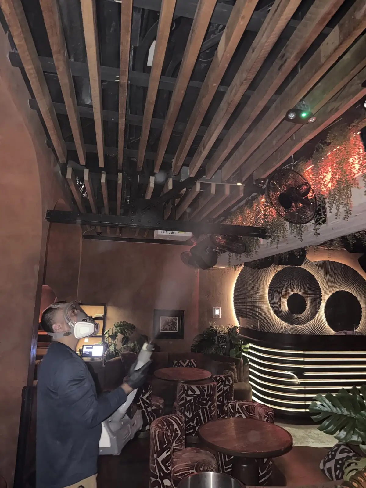 Technician in a full-face respirator fogging dining tables and a wood-slat ceiling in a Miami Beach restaurant.