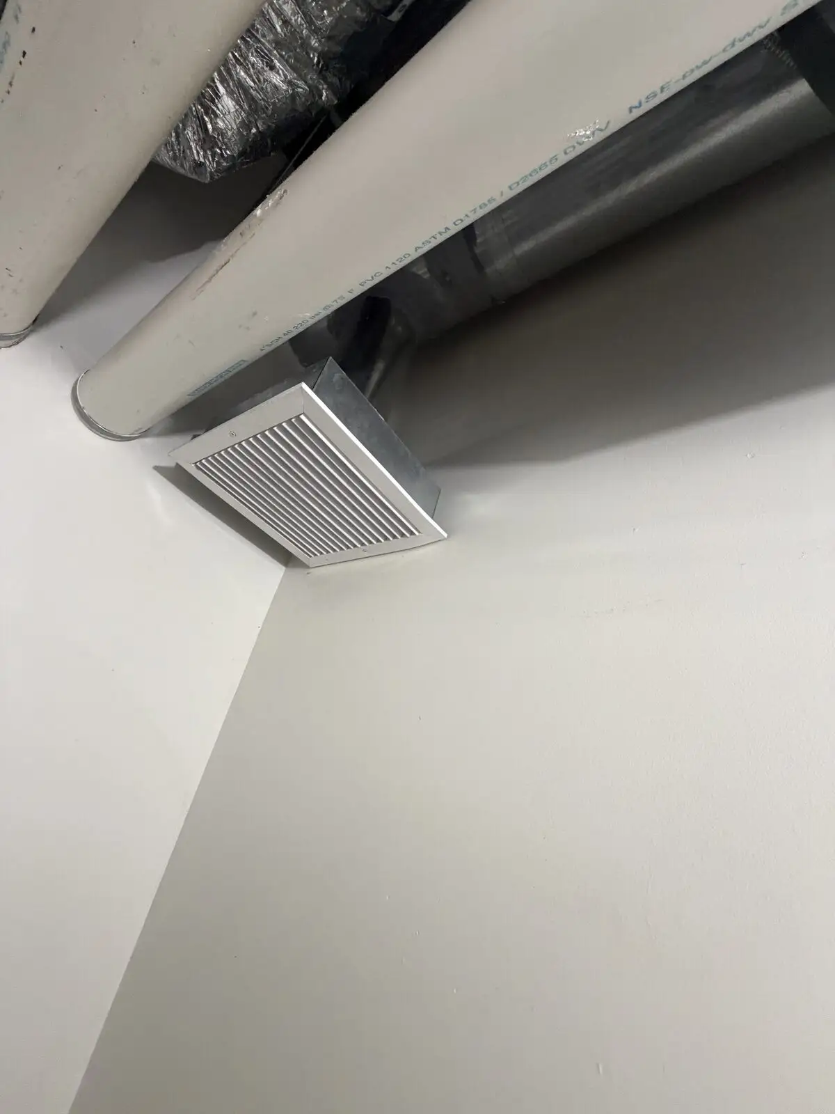 Clean white square HVAC vent in a corner below PVC and metal pipes in a Miami Beach restaurant kitchen.