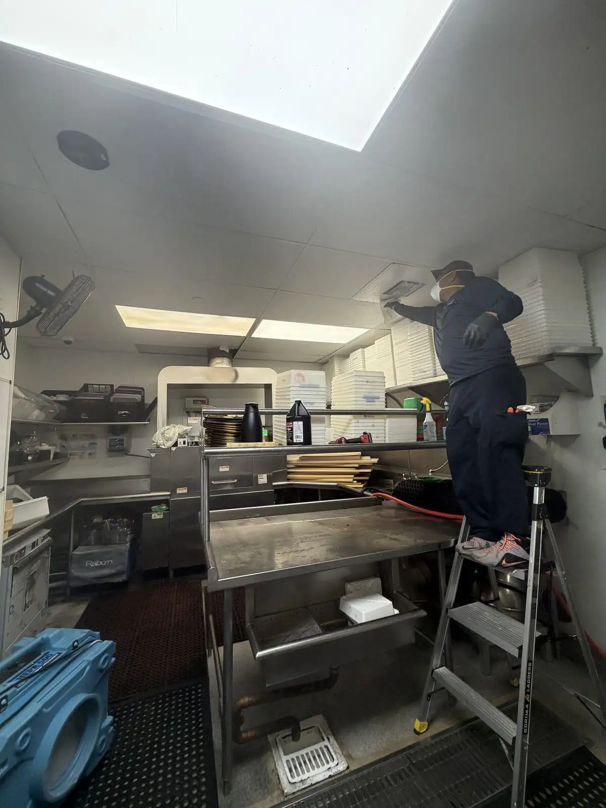 Technician in PPE on a step ladder wiping a kitchen ceiling HVAC diffuser above stainless prep tables and an air scrubber.