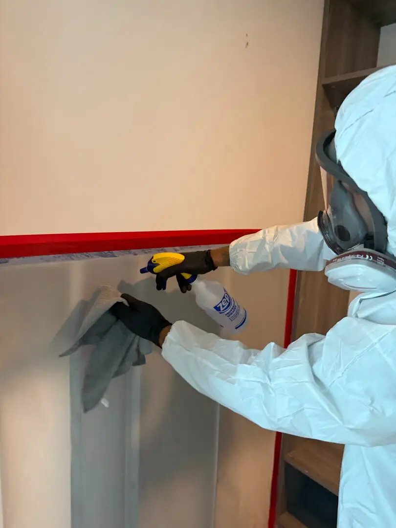 Technician in a hooded white suit and full-face respirator sprays and wipes a wall next to red-taped cabinetry.